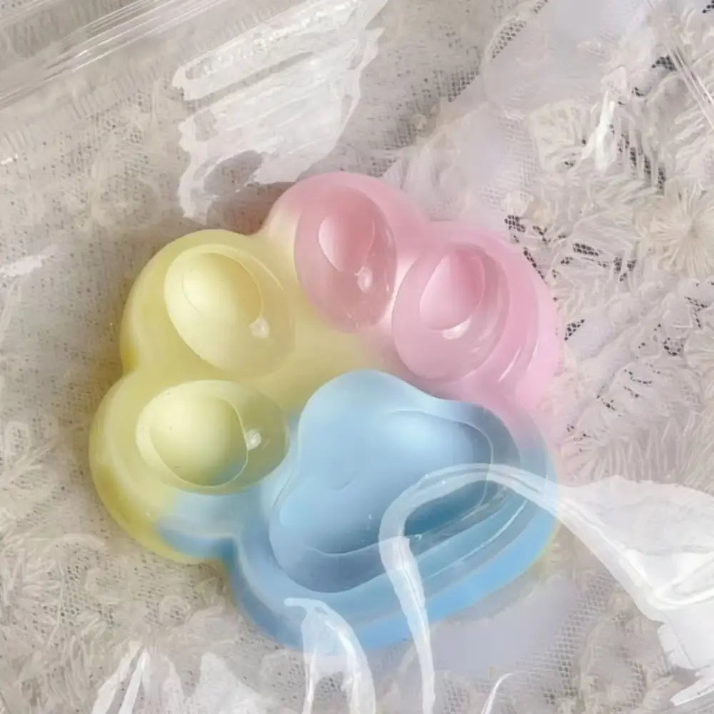 Jouet anti-stress souple et moelleux en silicone pastel, en forme de patte d’arc-en-ciel, rappelant des griffes de chat, avec des sections souples et translucides de couleur jaune, rose et bleue.