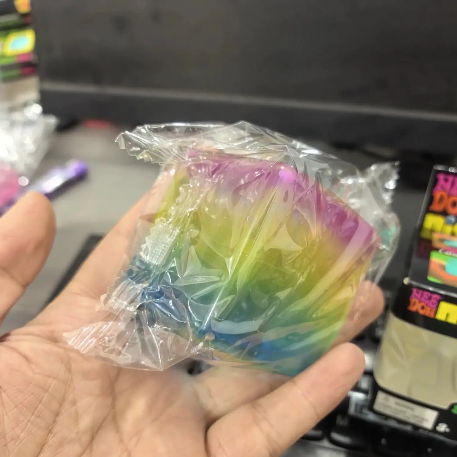 Eine Vorderansicht eines regenbogenfarbenen, transparenten Needoh-Würfel-Knet- und Fidget-Spielzeugs in klarer Kunststoffverpackung.