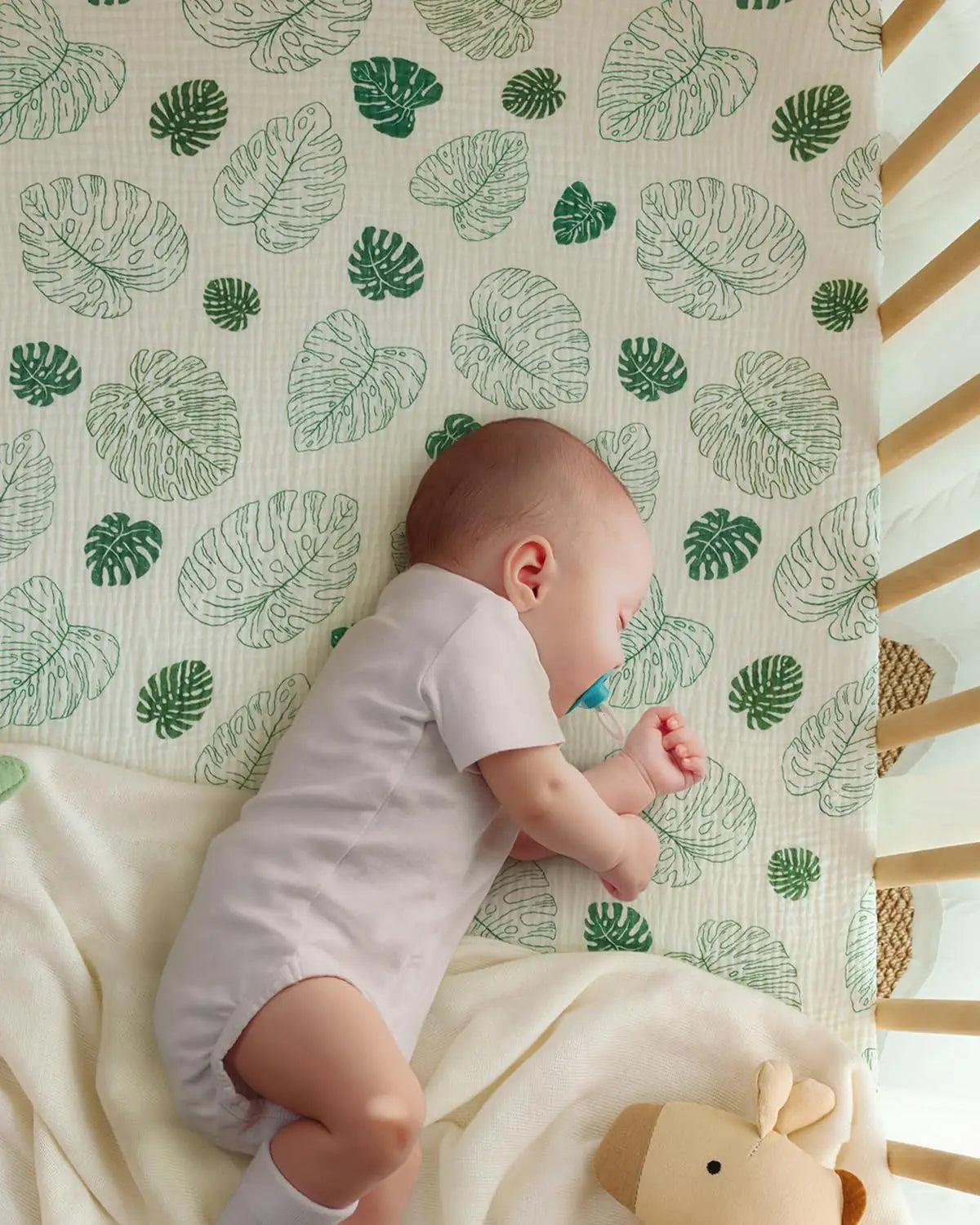 Ein Baby in einem weißen Body schläft seitlich auf einem Babybettlaken-Set aus cremefarbenem Baumwollmuslin mit grünen Monstera-Blattmustern.