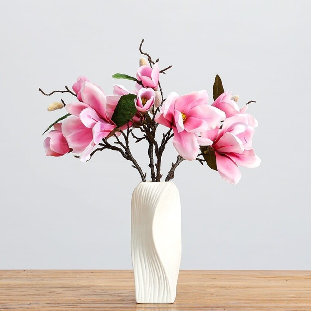 A white ceramic Nordic wave plastic flower vase with a sleek wavy design holds pink magnolia flowers and green leaves as aesthetic home decor.