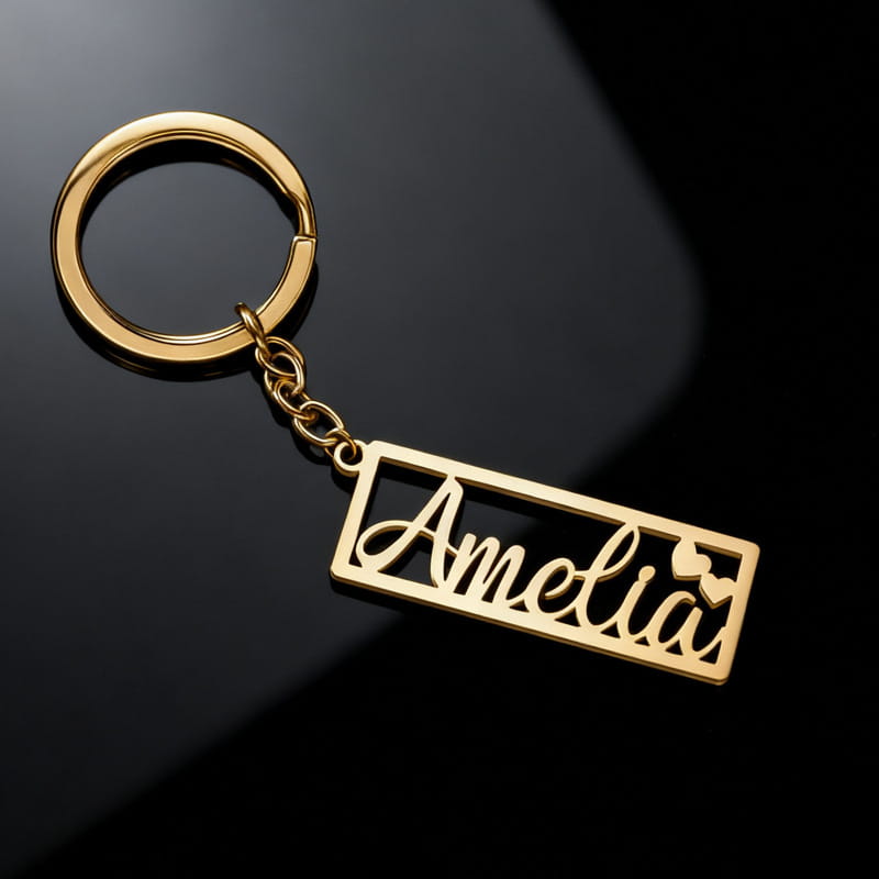Gold-colored stainless steel custom name keychain featuring the name amelia in elegant cursive script within a rectangular border accented by small heart cutouts.