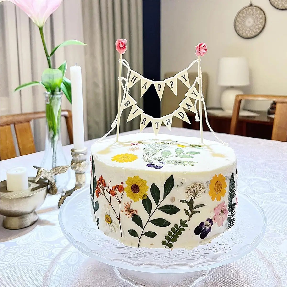 Un gâteau d’anniversaire blanc orné de fleurs pressées et de verdure, surmonté d’un fanion de gâteau d’anniversaire présentant des roses roses.