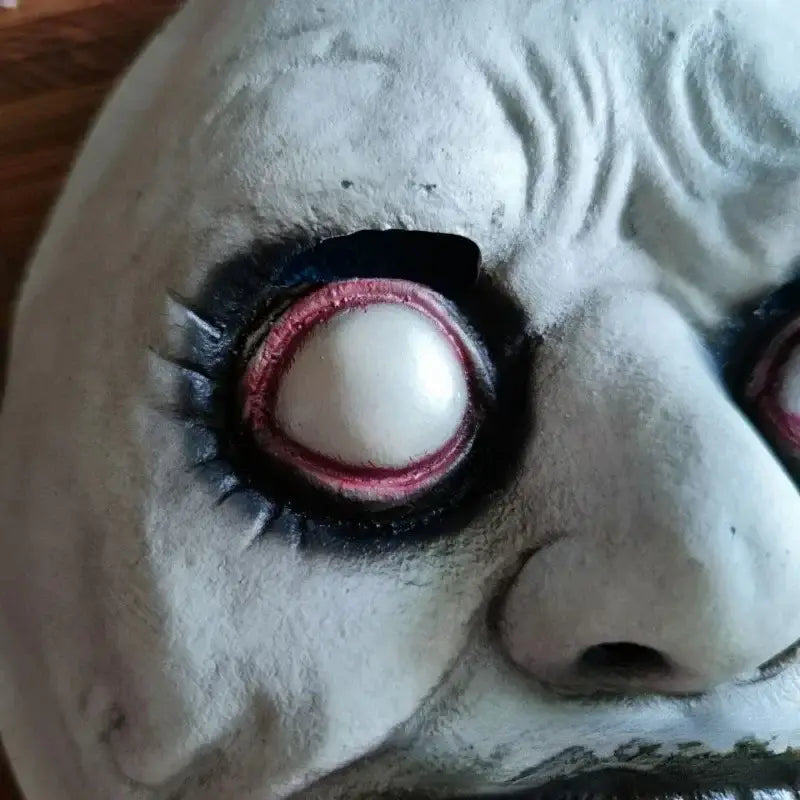 A close-up of a white textured resin creepy Halloween horror mask full face costume accessory with deeply set eyes featuring black eyelids and red-rimmed irises.