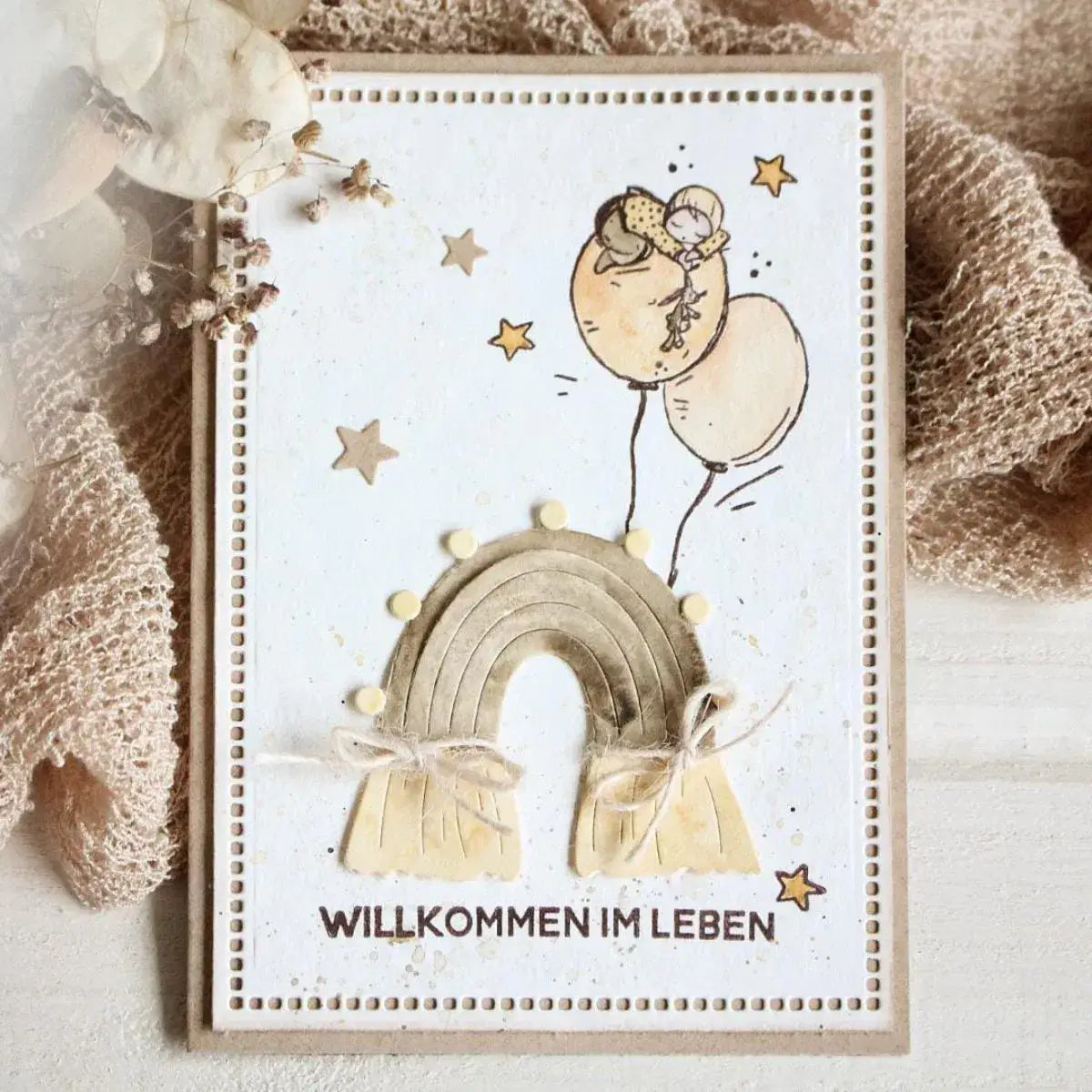eine handgefertigte Grußkarte mit einer verspielten Illustration eines Kindes auf Luftballons über einem strukturierten beigen Regenbogen mit kleinen gelben Pompons und Bindfäden; entworfen mit Oster-Ei-Hasen-Babyschneidvorlagen.