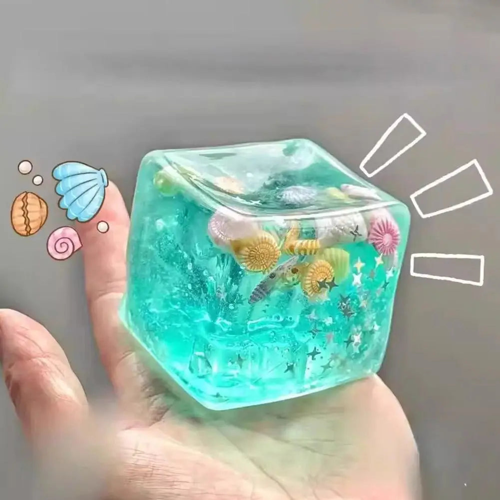 Un cube sensoriel à effet anti-stress turquoise translucide rempli de mini coquillages, d’étoiles de mer et de décorations scintillantes sur le thème de la mer, conçu comme un cadeau attentionné