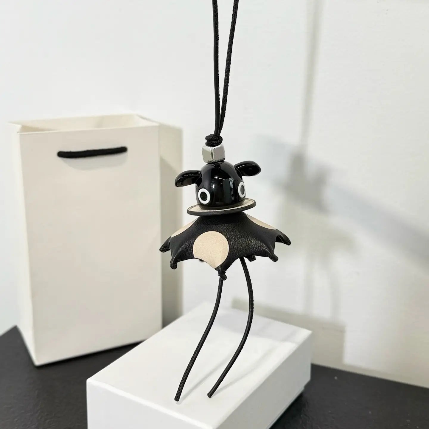 Llavero colgante para bolso con encanto de cuero con forma de murciélago negro, acabado brillante, con ojos circulares blancos y un parche de vientre color crema.