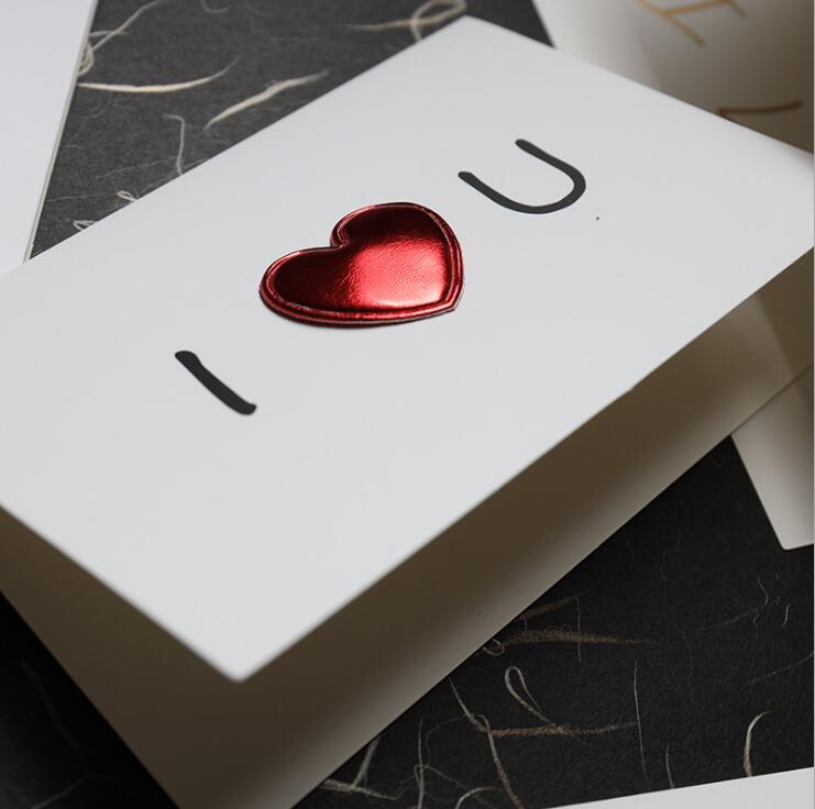 A glossy red heart-shaped charm made of metal affixed to a white red heart valentine greeting card wedding invitation with black i u lettering.