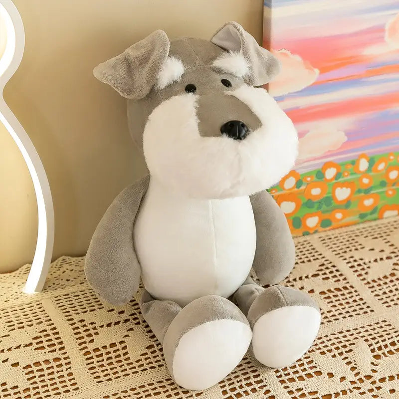 Un jouet de chien schnauzer en peluche gris et blanc, yeux et nez à boutons noirs, réalisé dans une matière souple, assis sur une nappe en dentelle, provenant de la collection de peluches d’animaux de la jungle et de poupées en peluche souples.