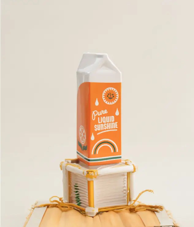 Vase en céramique représentant une brique de jus d’orange, de couleur orange, avec un design « pure liquid sunshine » comprenant un soleil souriant et un arc-en-ciel, un dessus en plastique blanc et une base rayée verte.