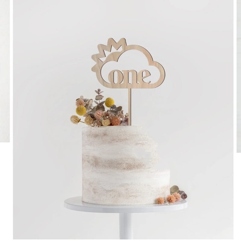 Un topper de gâteau en bois clair en forme de nuage, en bois, pour « one », portant le mot « one » découpé et surmonté de fleurs séchées, pour une décoration florale d’anniversaire.