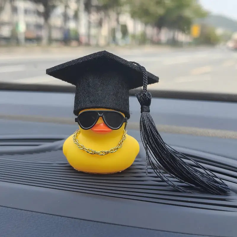 a yellow rubber duck graduation car dashboard ornament wearing a black graduation cap with tassel sunglasses and a gold chain necklace