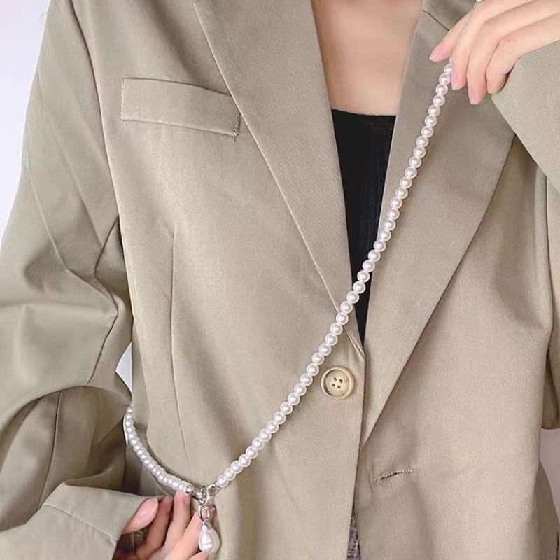 Un long collier délicat en perles en pendentif pour femmes, avec une bandoulière en chaîne de perles portée sur la poitrine d’un blazer beige.