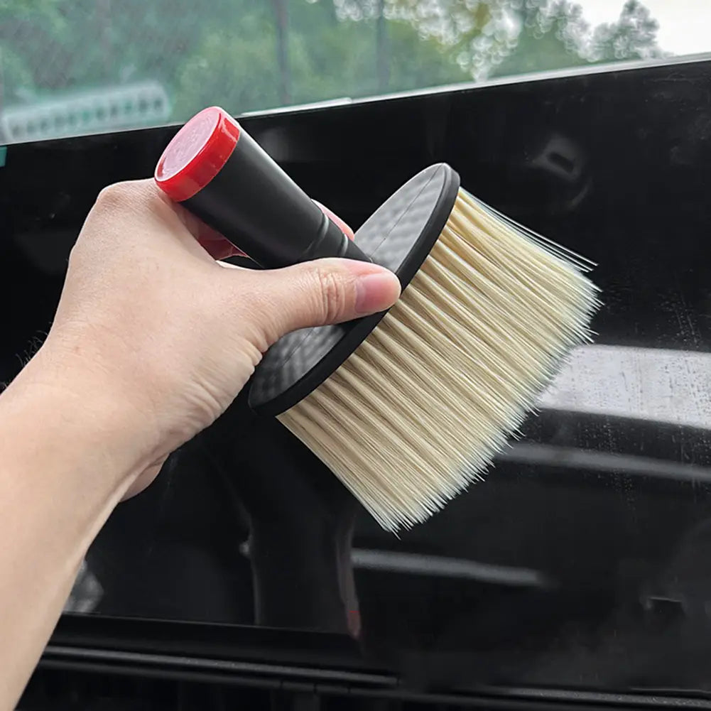 Eine Hand hält eine schwarze und rote Detailbürste für die Autopflege mit dichten hellbeigen Borsten; konzipiert als Werkzeug für eine schonende Innenraumreinigung.