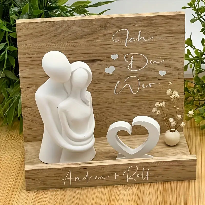 Statuette de couple en céramique blanche, aux formes arrondies et lisses, disposée sur une plaque en bois naturel gravée de texte romantique allemand et de noms, présentée comme un design de moule à bougie en silicone 3D où le couple s’embrasse.