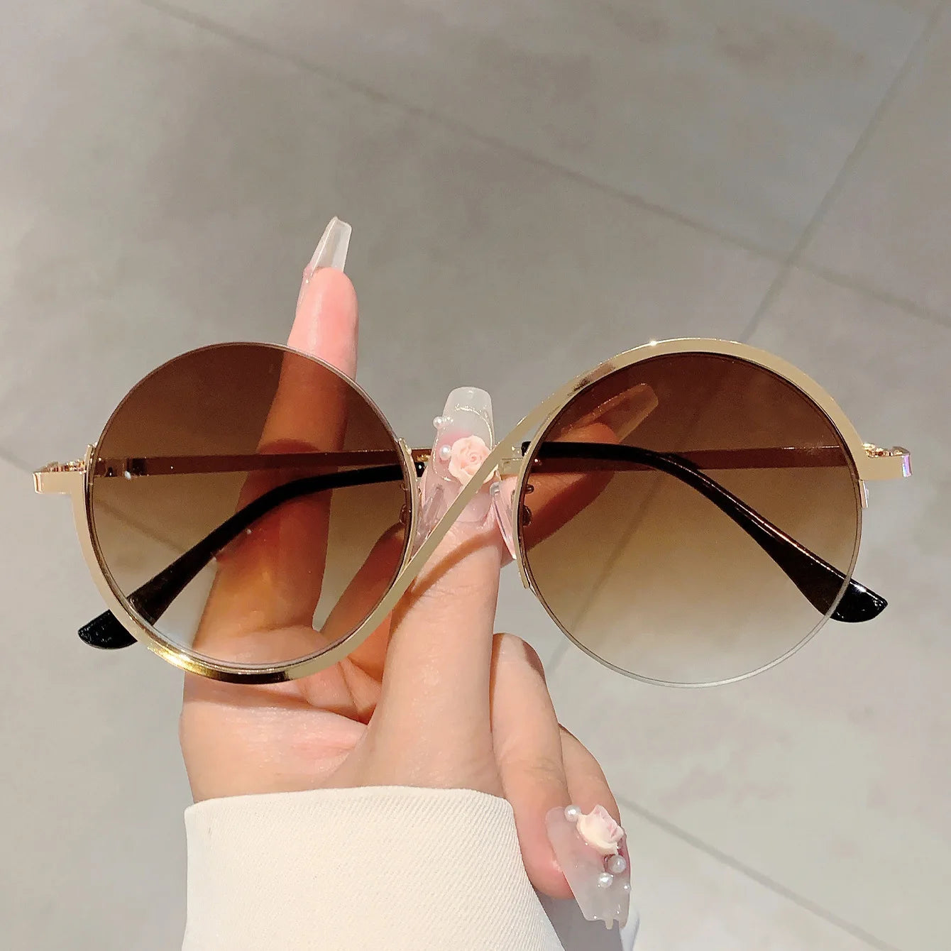 Unas gafas de sol con montura redonda con borde dorado, lentes marrones con degradado y puntas de patillas negras, sostenidas por una mano con arte de uñas decorativo; adecuadas para atuendos de moda y diversas situaciones aplicables.