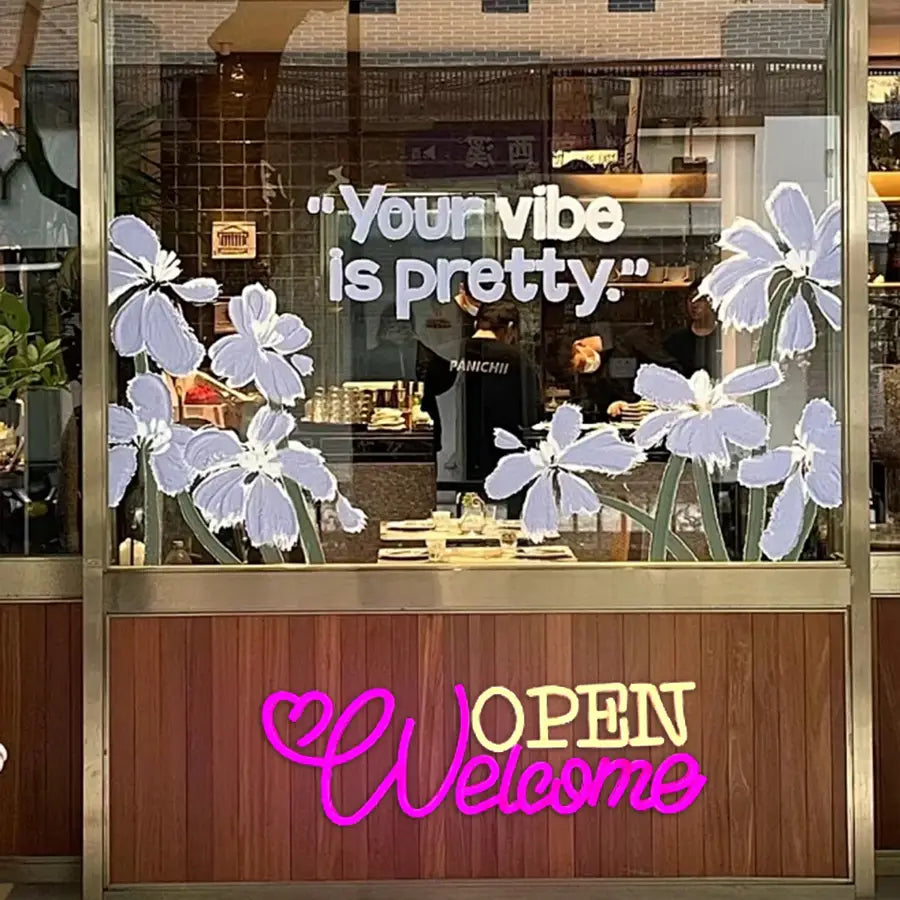 La vitrine présente une enseigne au néon à LED ouverte, décoration murale alimentée par USB, avec un décor floral de fleurs de couleur lavande et de centres blancs, ainsi que des tiges vertes autour de la phrase « Votre vibe est jolie ».
