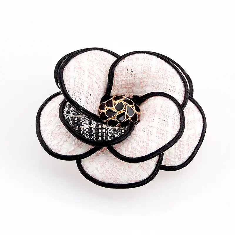 Zarte Brosche aus weißem und schwarzem Kamelienstoff in Form einer Blume mit strukturierten Blütenblättern und einem goldfarbenen Mittelteil, verziert mit schwarzen Emaille-Details, entworfen als Retro-Damenbrosche.
