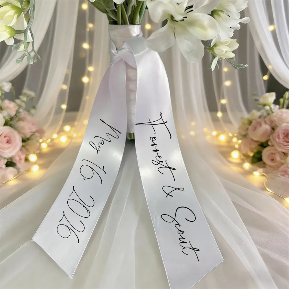 Un ruban en satin blanc avec un lettrage cursif noir indiquant « forever » et « scout », ainsi qu’un ruban de bouquet de mariage personnalisé avec « may 16 », portant les noms propres et la date