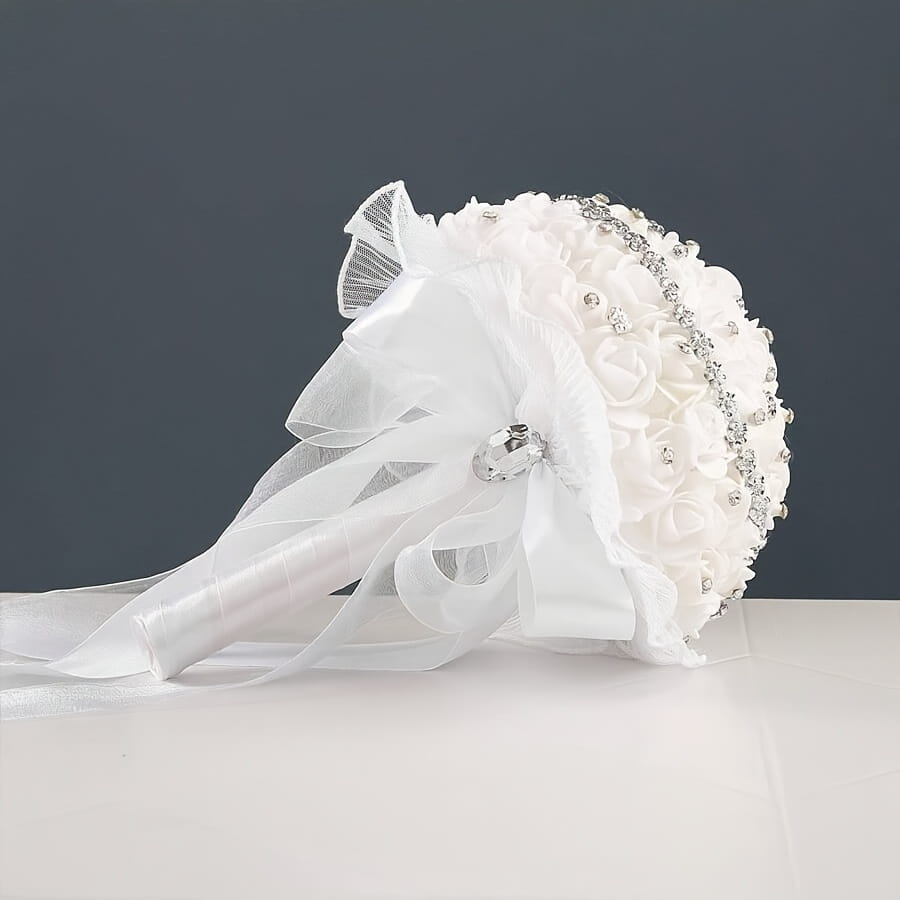 White bridal bouquet with rhinestones and lace featuring delicate fabric roses sparkling rhinestone embellishments and a sheer ribbon bow tied around its handle.