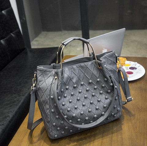 Bolso de hombro de cuero gris con tachuelas, textura acolchada y herrajes metálicos que muestran el compartimento interior.