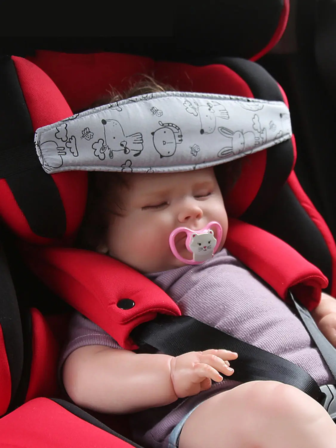 Ein weicher grauer verstellbarer Kindwagensitz-Kopflehnengurt-Polster mit schwarzen Cartoon-Tiermotiven, das über die Stirn des Babys in einem roten und schwarzen Autositz zur Schlafpositionierung gelegt ist.