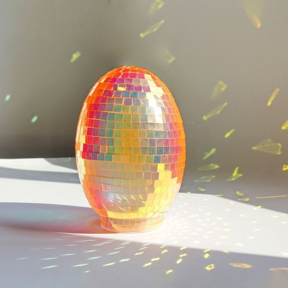 An iridescent glass disco egg ball ornament featuring a vibrant egg-shaped design with reflective mosaic tiles in shades of orange pink and gold.