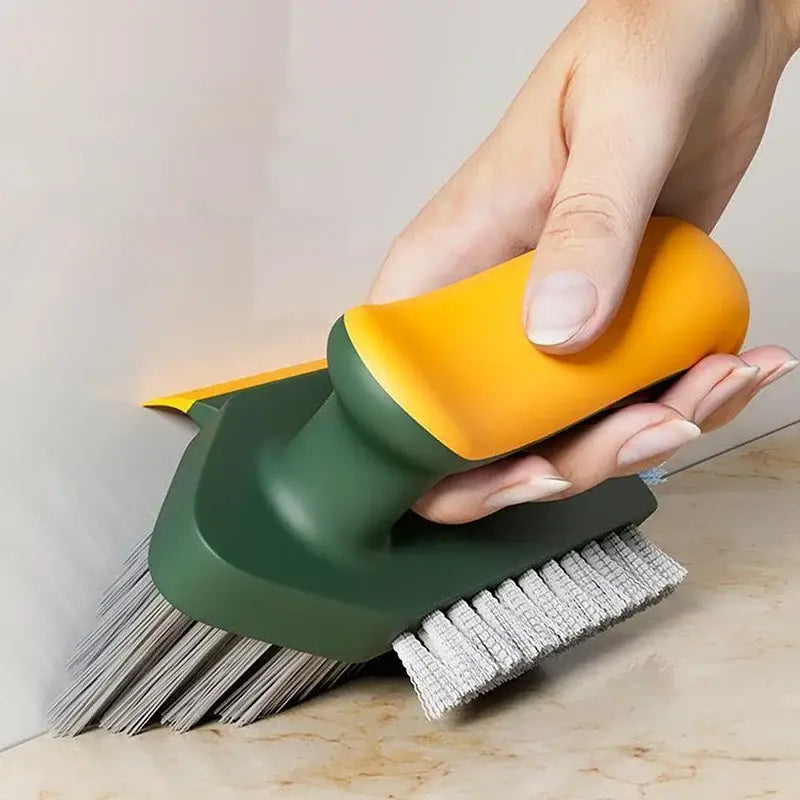 Une main tient une brosse de nettoyage en forme de V pour carrelage et rainures, ainsi qu’une brosse à crevices, avec un manche en plastique orange et vert doté de poils métalliques rigides pour le récurage et de poils en tissu gris plus souple pour un nettoyage délicat.