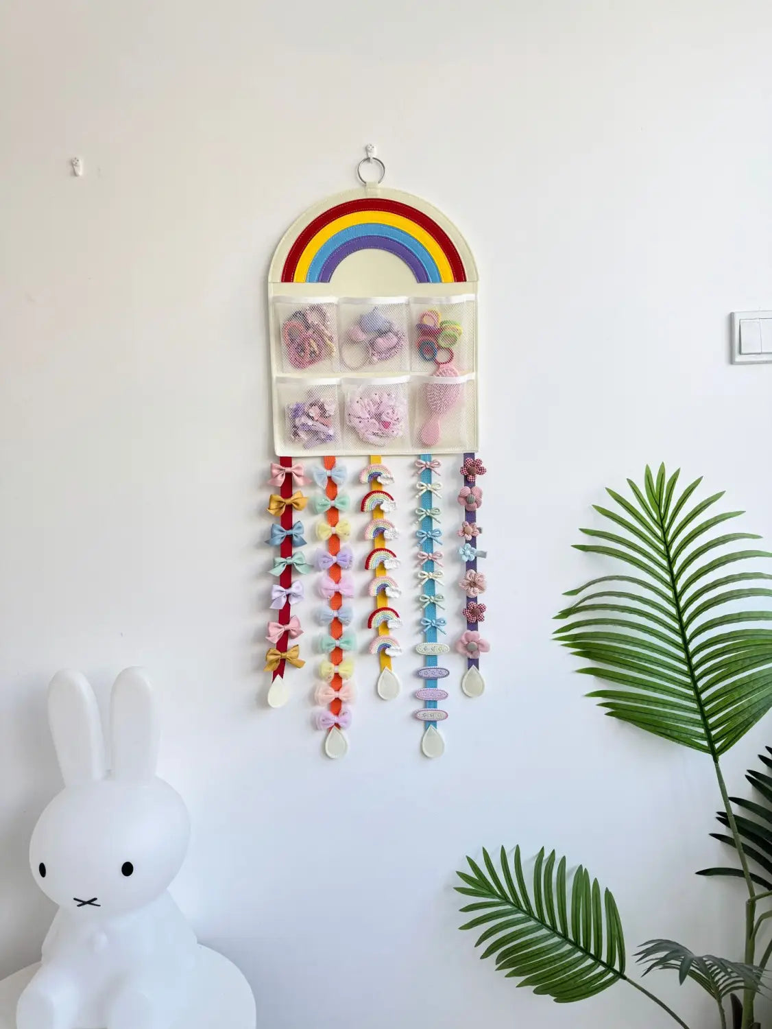 Organizador colgante para accesorios de cabello con temática de arcoíris, de pared, con bolsillos de tela coloridos y cintas decoradas con lazos colgantes.
