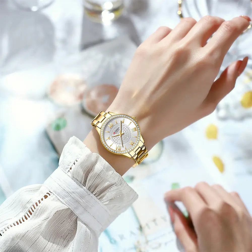 Curren women stainless steel crystal quartz watch with a gold-toned band, sparkling diamond-encrusted bezel, and white dial featuring roman numerals.
