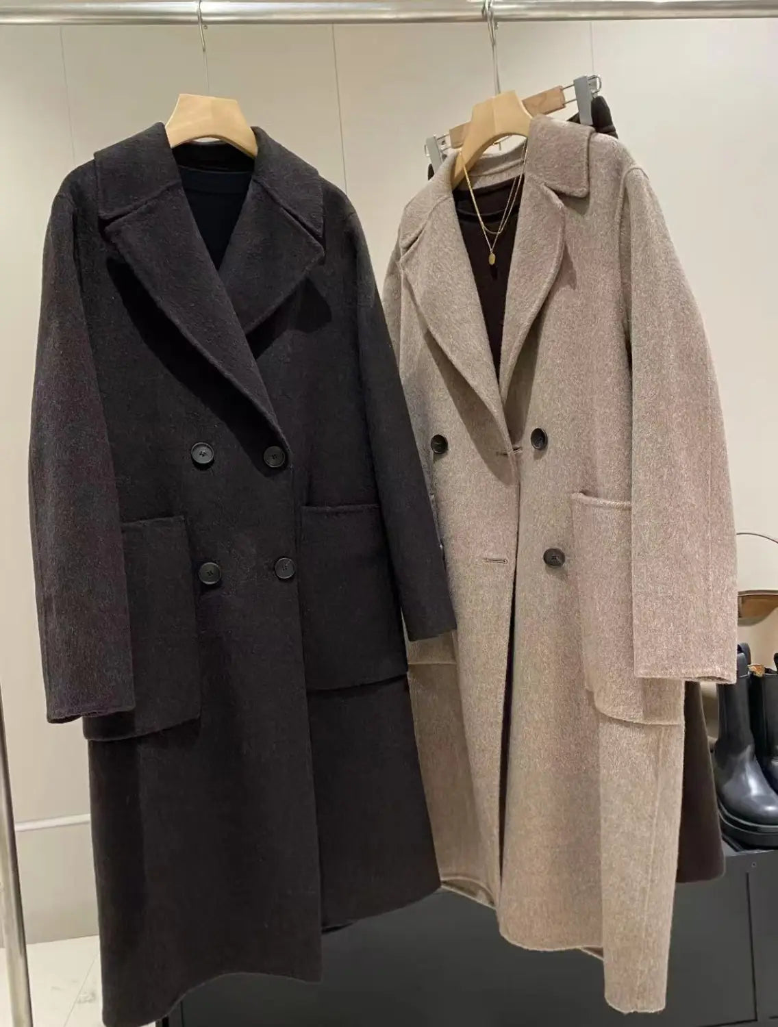 Two wool blend double breasted coats in dark charcoal and beige hang side by side on wooden hangers.