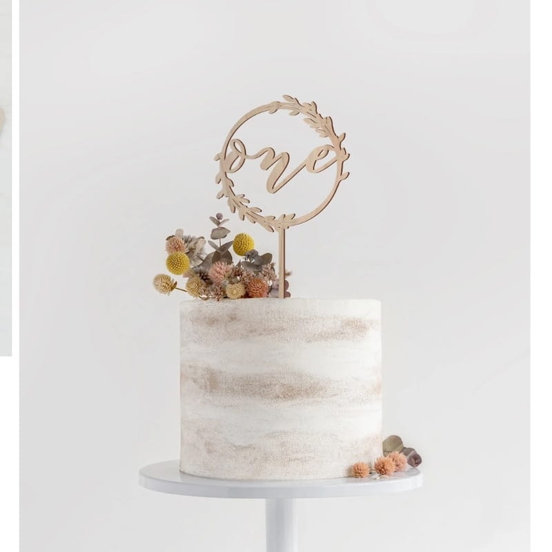 Un délicat topper de gâteau en bois, pour « one », de couleur naturelle, présentant le mot « one » inscrit avec élégance dans un motif de couronne circulaire, orné de fleurs séchées et de baies, pour une décoration florale d’anniversaire.