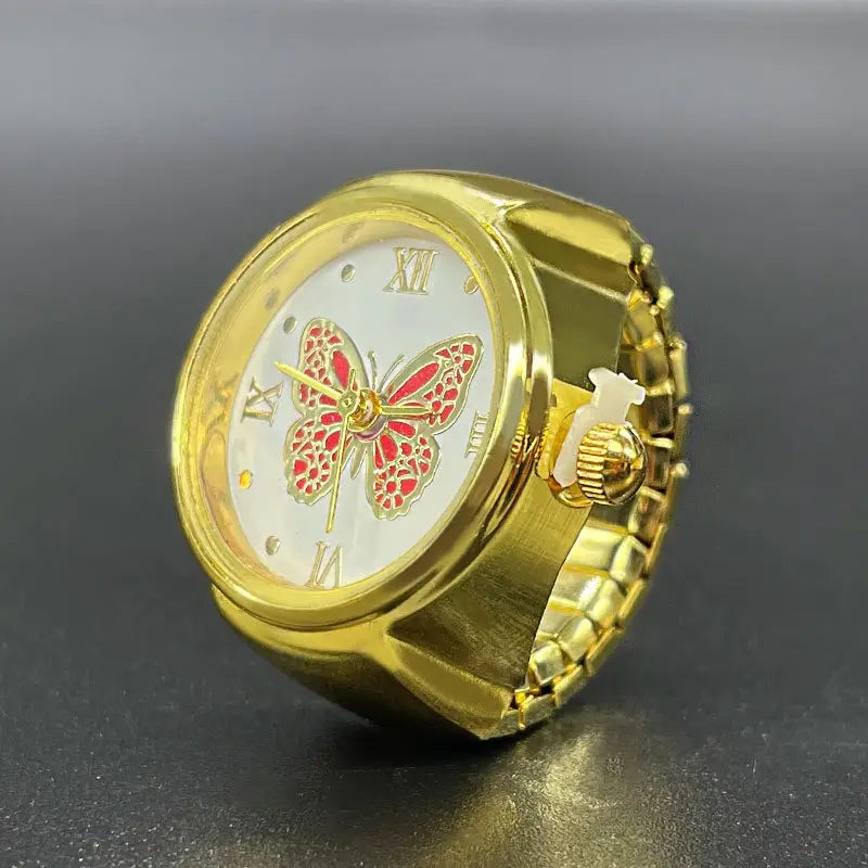 Gold-toned stainless steel ring-style watch with a white face featuring roman numerals and an ornate red butterfly design, a cute piece of jewelry.