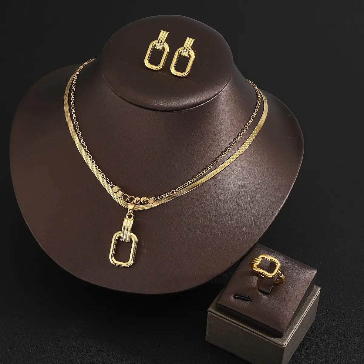 Juego de joyería de acero inoxidable color dorado para mujeres, con un collar de doble capa con cadena de acabado dorado, colgante de forma ovalada y tres barras verticales mostradas en un maniquí de cuello color marrón.