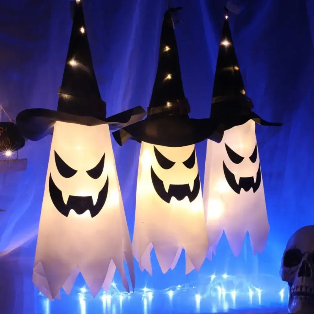 Décoration d’Halloween suspendue en forme de fantôme à DEL, comprenant trois lanternes blanches lumineuses avec des chapeaux de sorcière noirs et des expressions faciales sculptées sur un fond bleu.
