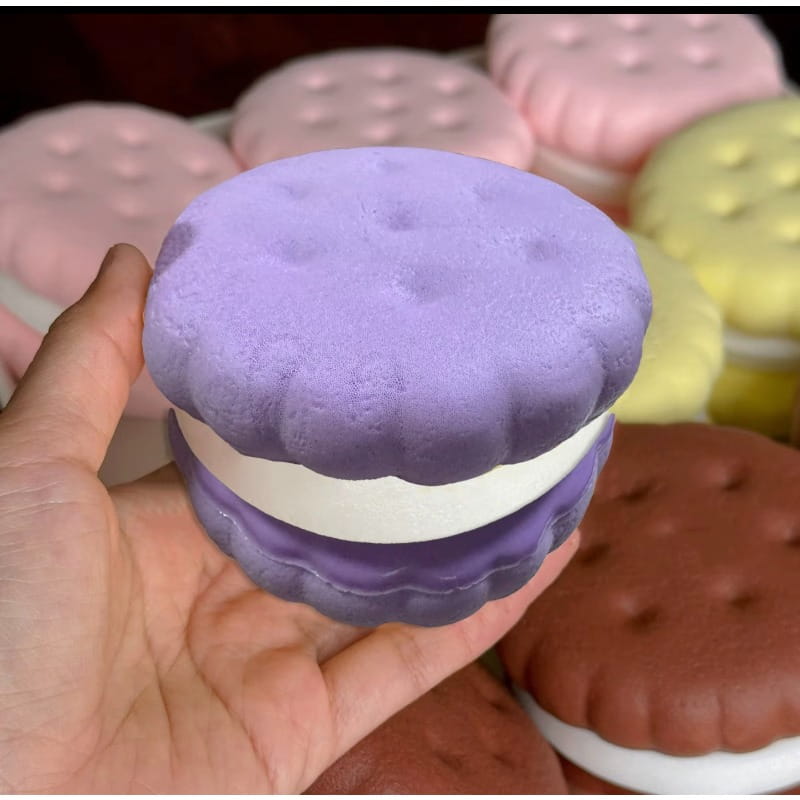 Eine Hand, die ein lila-weißes, langsam ansteigendes großes Keks-Knautschspielzeug hält, geformt wie ein Eiscreme-Sandwich mit realistischer Kuchenstruktur.