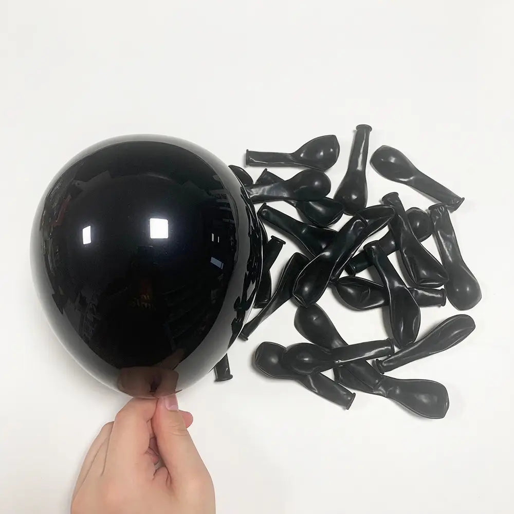 Ein glänzender schwarzer Latexballon, der von einer Hand gehalten wird, mit mehreren entfalteten schwarzen Latexballons, die in der Nähe verstreut sind, als Teil eines roten-schwarzen Ballon-Girlandenbogen-Sets zur Partydekoration.