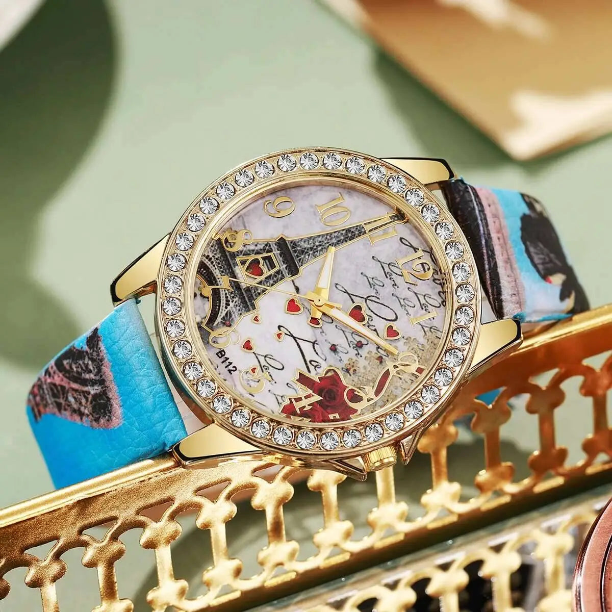 A gold-toned eiffel tower floral quartz women watch with a sparkling crystal bezel and an ornate face featuring playing card motifs on a light background.