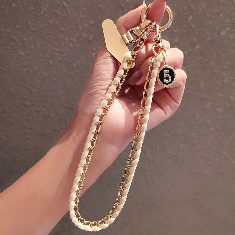 Ein cremefarbener Handy-Handgelenkriemen aus goldfarbener Kette als Anti-Lost-Handylanyard, mit goldfarbenen Metallgliedern und einem runden Anhänger mit der Zahl 5 in Schwarz, verziert mit funkelnden Strasssteinen.