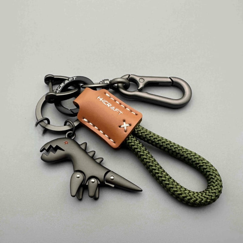 llavero metálico con forma de dinosaurio de color gris oscuro, con etiqueta de cuero marrón y cuerda trenzada verde oliva para un llavero metálico de cuero con forma de dinosaurio trenzado duradero.