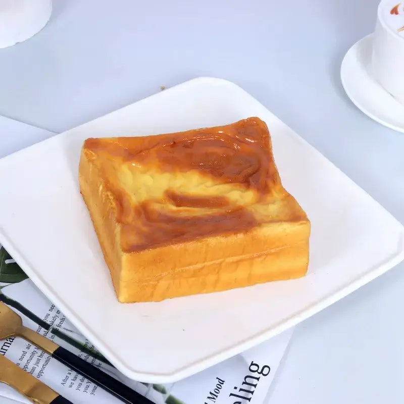 Design einer goldbraunen, glänzend marmorierten Kuchenscheibe auf einem Hundebrot – langsam aufsteigendem, weichem Knetspielzeug, dargestellt auf einem weißen quadratischen Teller
