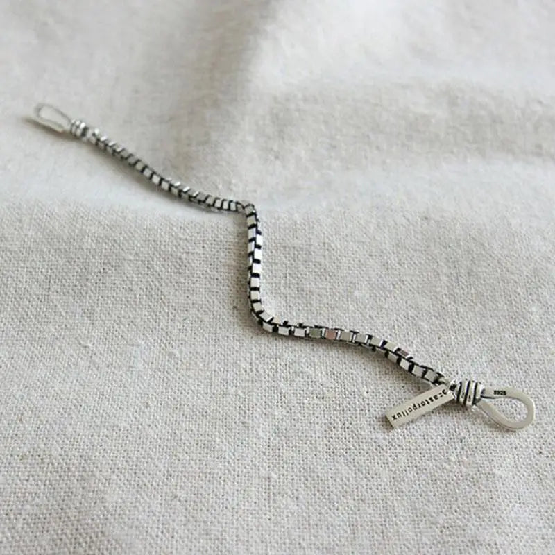 L’image présente un bracelet de charme en métal géométrique en chaîne ton argent pour couples, avec un motif caractéristique à carreaux noir et blanc et une petite plaquette rectangulaire gravée, à l’aspect vieilli, près de la fermeture.