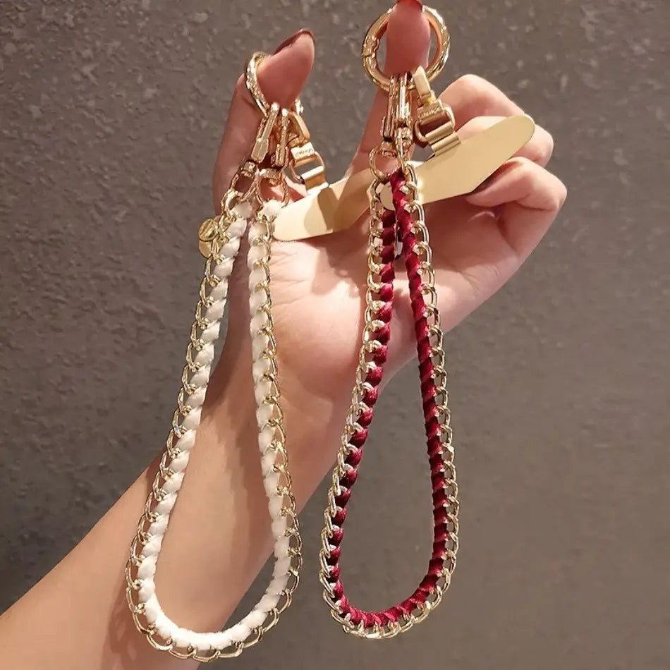 Eine Hand hält zwei Handy-Handgelenkriemen aus goldfarbener Kette als Anti-Lost-Handylanyards in Weiß und Rot, mit geflochtenen Korden, die mit goldfarbenen Metallketten umwickelt sind, sowie passenden metallischen Verschlüssen.