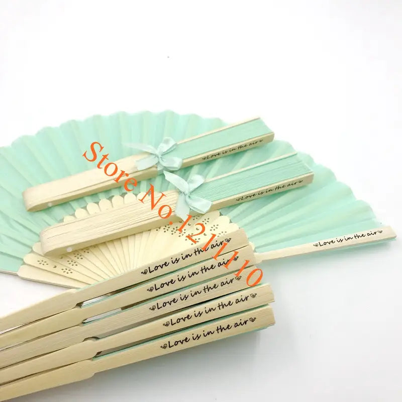Pastel mint and cream-colored custom wedding hand fans made of personalized bamboo silk with delicate lace detailing and love is in the air engraved on each.