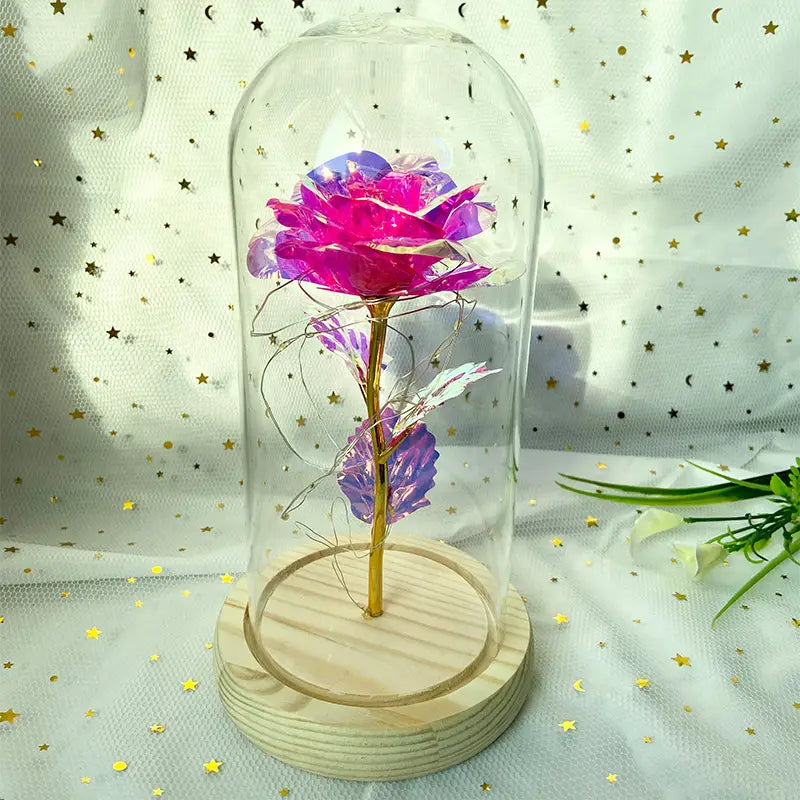 Eine kräftig pinke und violette metallische Rose mit goldenem Stiel, umschlossen in einer klaren Glaskuppel auf einem hellen Holzsockel, im „Eternal Gold Rose Glass Dome Led Light Gift“.