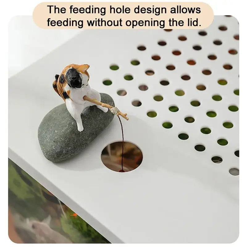 cute calico cat figurine perched on a smooth gray stone holding a fishing rod with line extending through a circular feeding hole in the white lid of a transparent desktop fish tank.