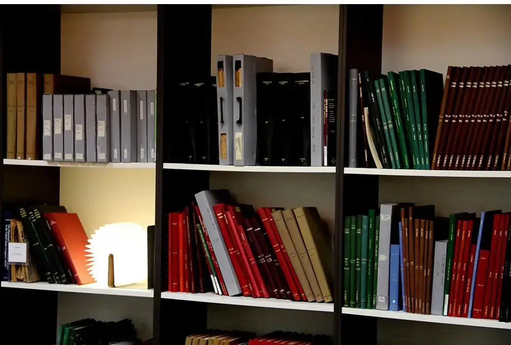Lámpara de mesa recargable con luz LED plegable con forma de libro colocada sobre una estantería de madera oscura con estantes blancos, con libros multicolores organizados cuidadosamente, vista desde el ángulo frontal.