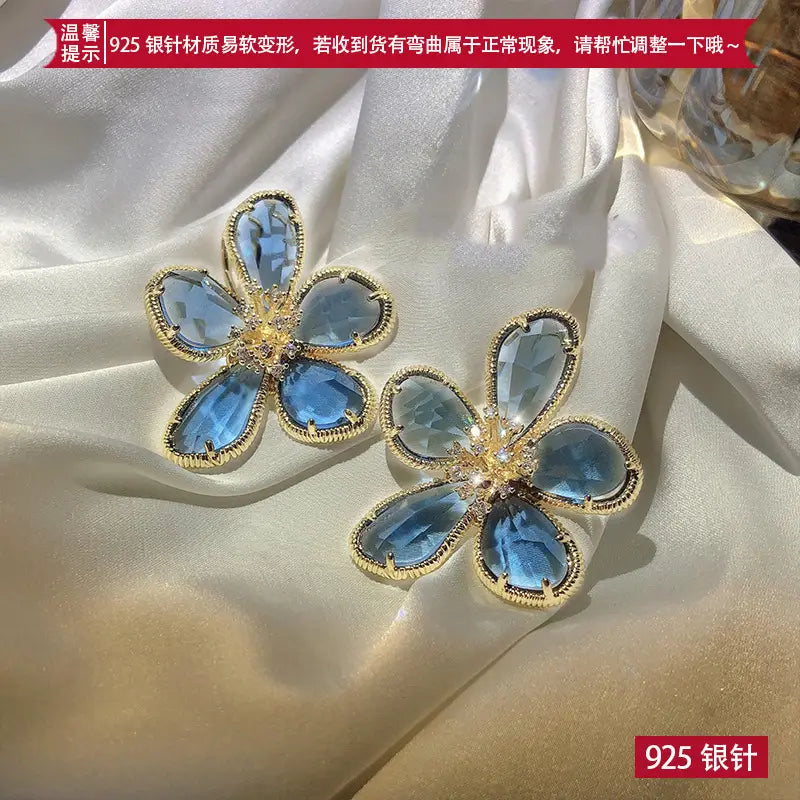 金色調の金属フレームを用いた、エレガントな青い花形のファセットカット宝石を特徴とするゴールドフラワークリスタルスタッドピアスの1ペア。