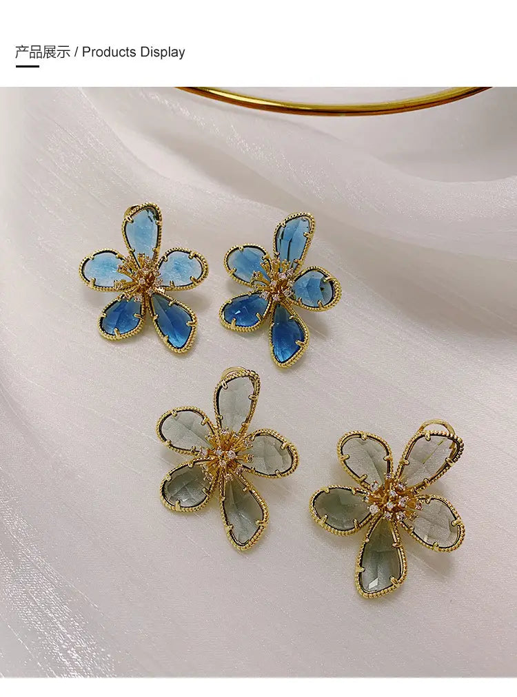 金色調の金属フレームで仕立てた、繊細な半透明のブルーおよびグリーンのガラスの花びらを特徴とするゴールドフラワークリスタルスタッドピアスの1ペア。