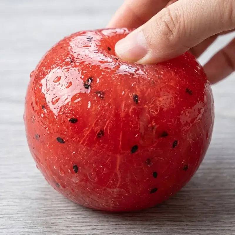 A glossy red watermelon-shaped squishy stress relief toy with black seeds embedded in its surface.
