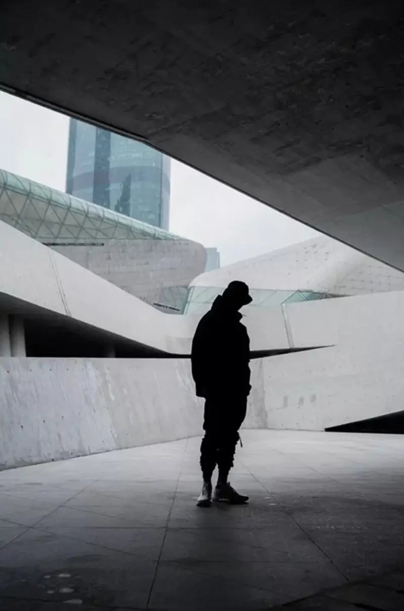Silhouette d’une personne portant une veste imperméable à capuche tactique techwear noire, vue de dos, sous une architecture urbaine en béton moderne.